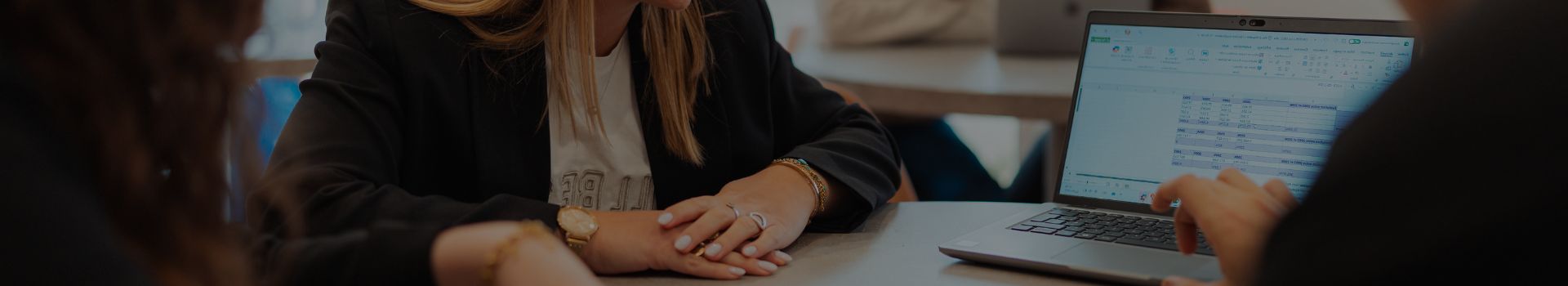 Les mains de trois étudiants assis autour d'une table avec un ordinateur dessus, une main pointe du doigt l'ordinateur qui est face caméra