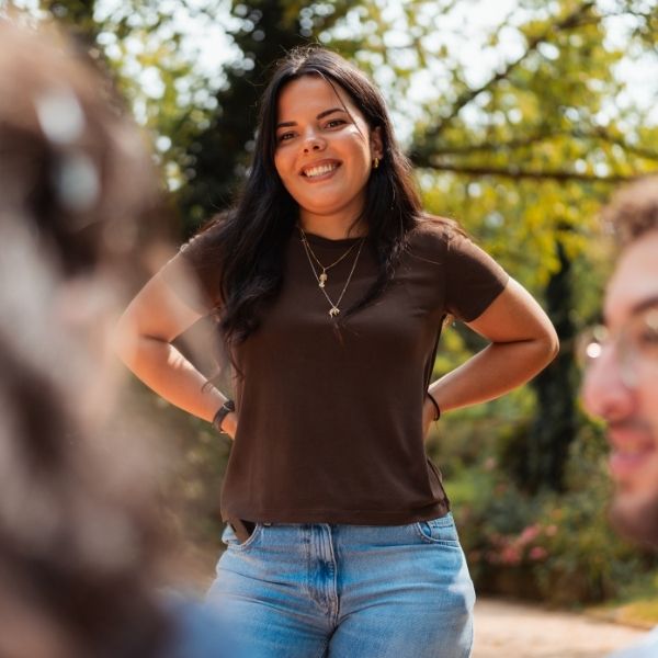 Jeune femme brune en t-shirt marron et jean bleu qui sourit dans un parc