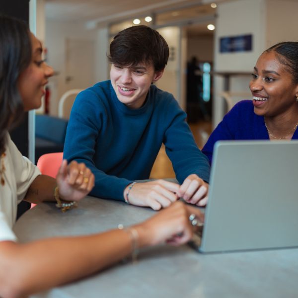 Trois étudiants assis autour d'une table avec un ordinateur dessus et discutent