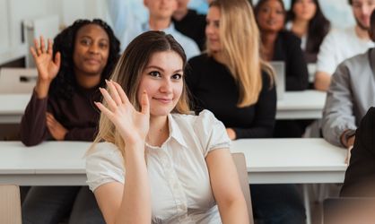 Jeune femme blonde avec une chemise blanche lève la main dans une salle de classe