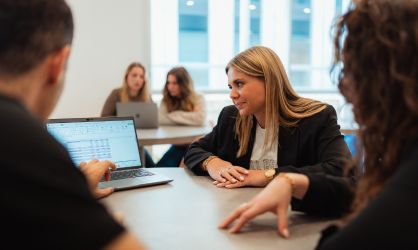 Trois étudiants assis autour d'une table avec un ordinateur dessus, une main pointe du doigt l'ordinateur qui est face caméra